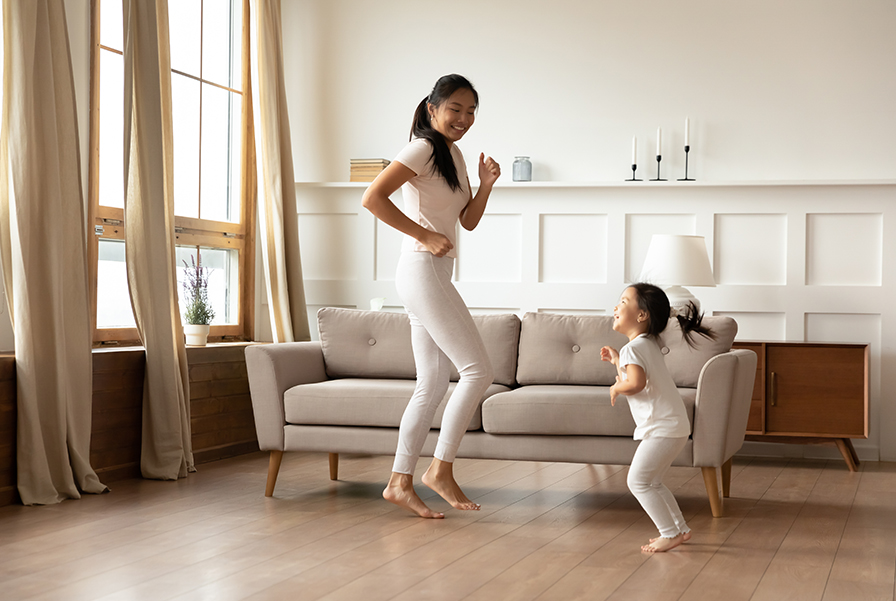 A smiling mother and her young daughter playfully dance in a modern, sunlit living room with a beige sofa. This image represents the personal, functional, and inviting interior spaces that can be designed with an interior architecture degree.