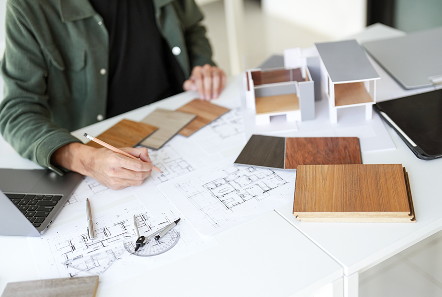A young professional works on architectural blueprints and a 3D model of a house, surrounded by wooden material samples and design tools. This image highlights the creative and technical skills—from drafting to material selection—gained through an interior architecture program.