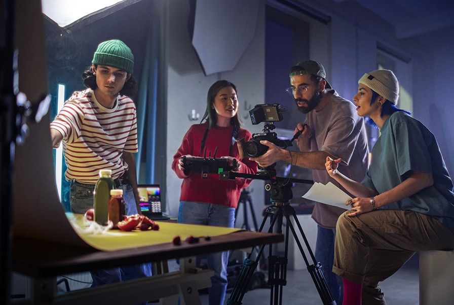 A small production crew discusses a shot around a table with a yellow backdrop, where bottles and fruit are placed and are using a professional video camera on a tripod and studio lighting.