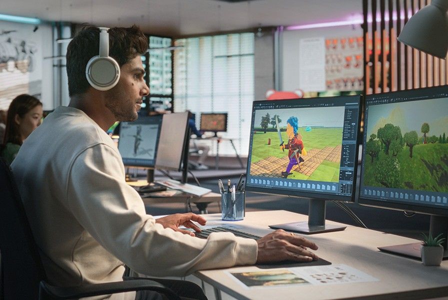 A man with white headphones sitting at a desk while working on a game design on a monitor screen.