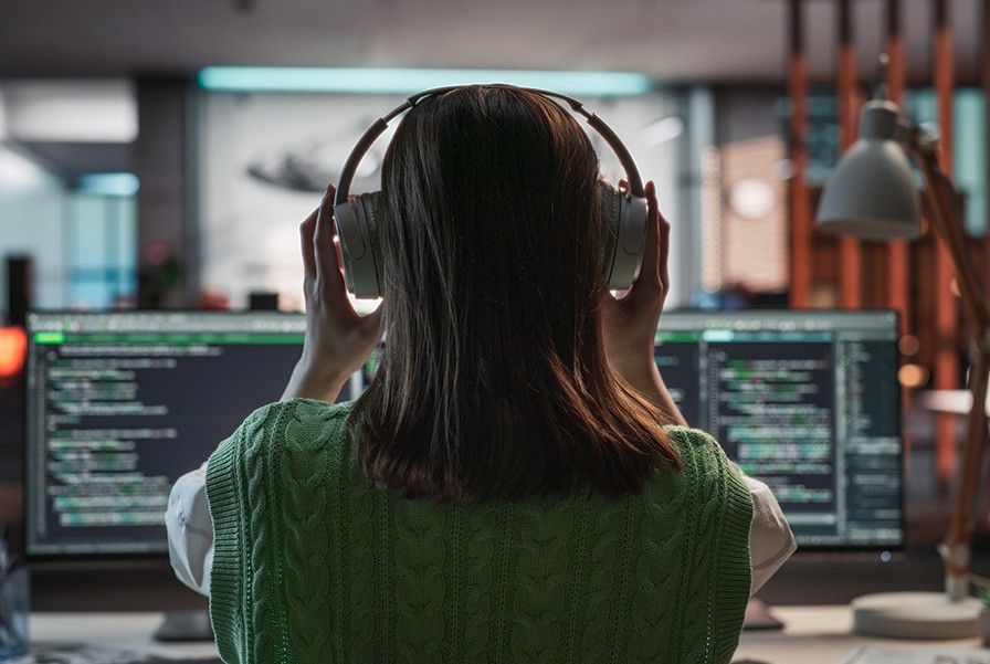A back view of a woman touching her headphones that she has on while sitting in front of gaming code sources which are on the monitor screens.