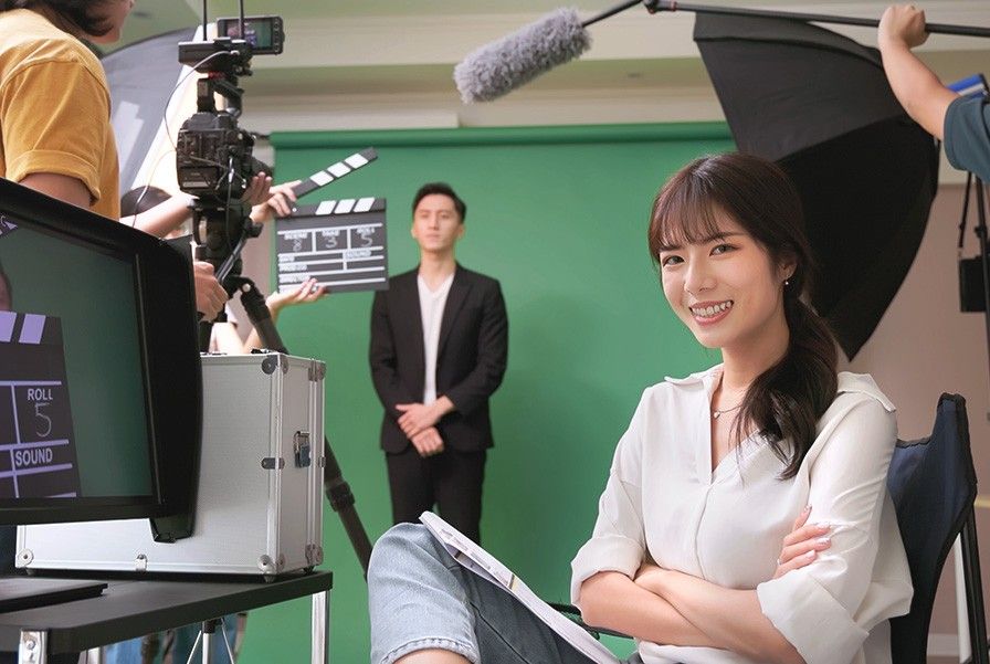 A woman in white blouse smiling while sitting in front of a recording setting where a man in blazer is standing in front of a green screen with a camera and microphone in front of him.
