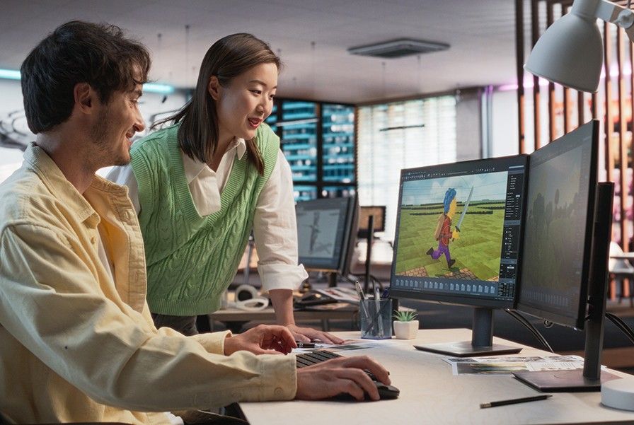 Two game developers review a 3D character model on a dual-monitor computer in a modern office.
