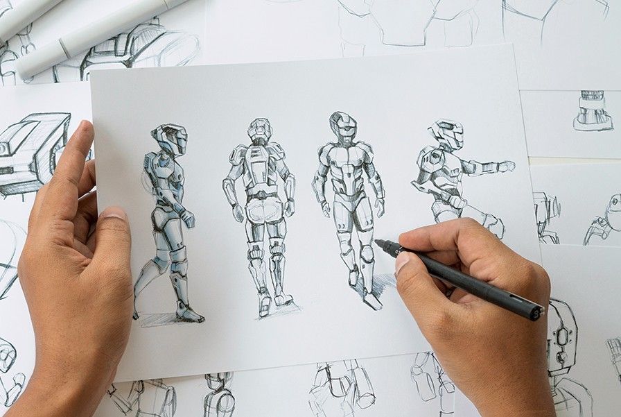 A person's hands drawing four sketches of a futuristic robot character on paper using a pen above a pile of other robot sketches.
